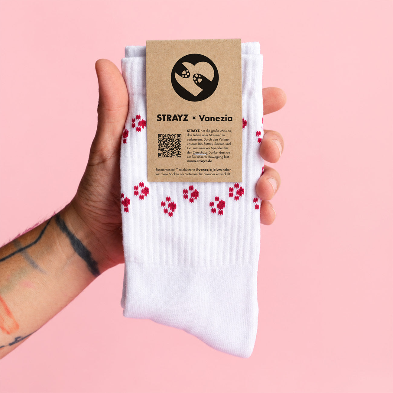 Hand hält ein Paar weiße STRAYZ × Vanezia Socken mit roten Pfotenabdrücken und Banderole aus Kraftpapier vor rosa Hintergrund