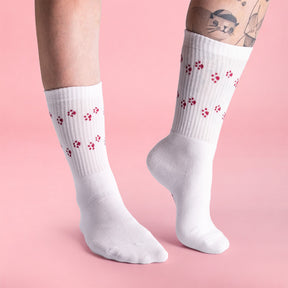 Person trägt weiße STRAYZ × Vanezia Socken mit roten Pfotenabdrücken, am Bein ein Tattoo einer Katze, vor rosa Hintergrund