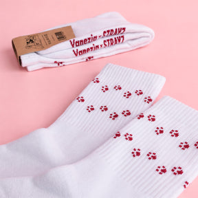 Weiße STRAYZ × Vanezia Socken mit roten Pfotenabdrücken liegen auf rosa Untergrund, dahinter ein gefaltetes Paar mit rotem Schriftzug "Vanezia × STRAYZ"
